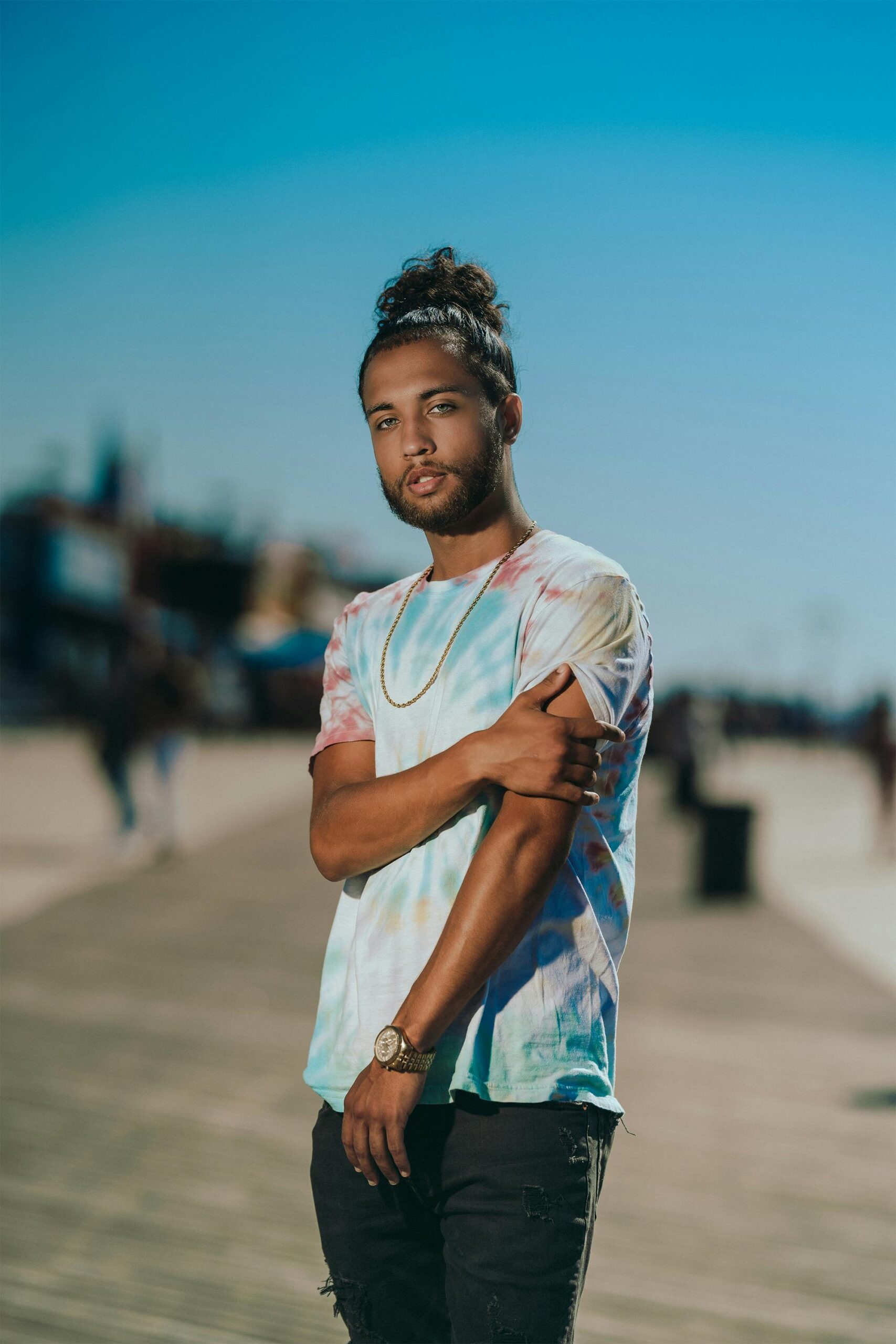 A fashionable adult male stands confidently on a sunny boardwalk in New York.